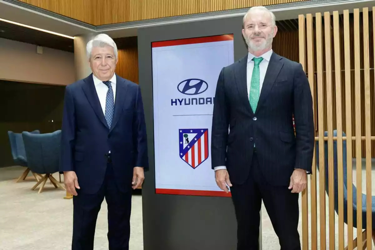 Dos hombres de traje posan junto a una pantalla que muestra los logotipos de Hyundai y el Atlético de Madrid en una sala moderna Dos hombres de traje posan junto a una pantalla que muestra los logotipos de Hyundai y el Atlético de Madrid en una sala moderna