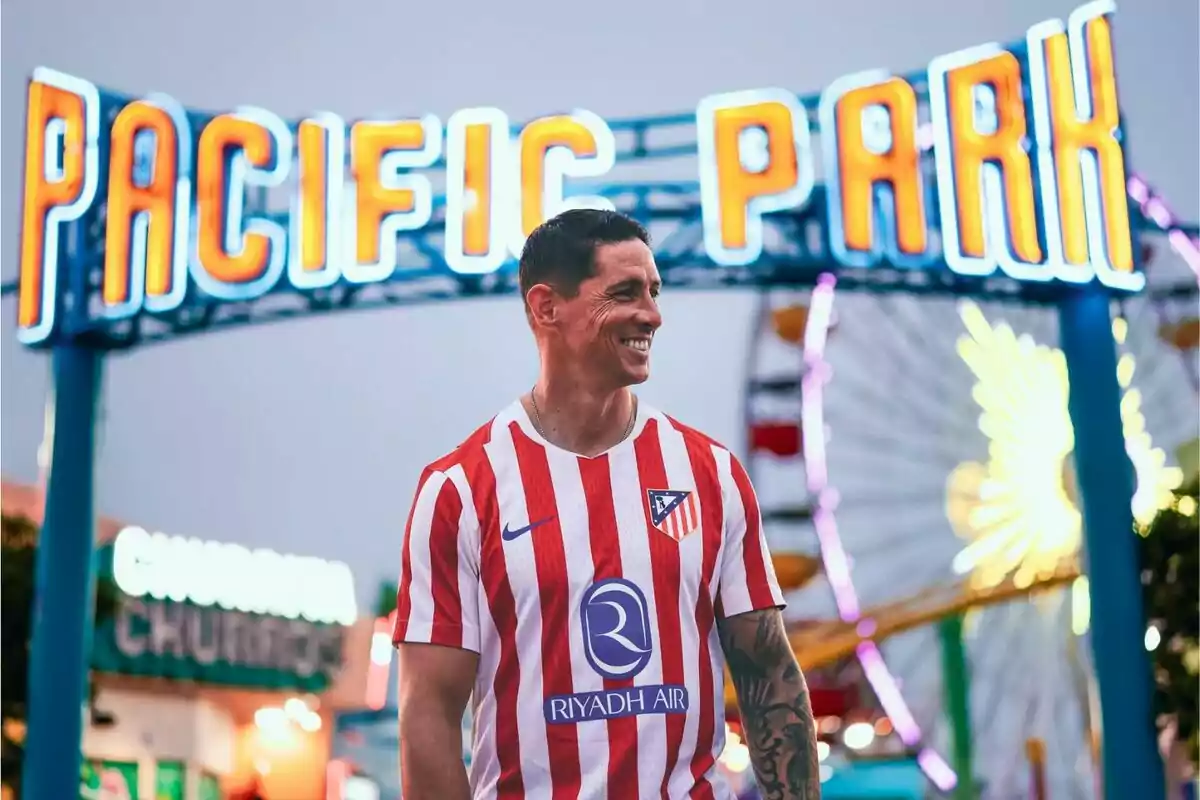 Hombre sonriente con camiseta del Atlético de Madrid posando frente al letrero iluminado de Pacific Park y una rueda de la fortuna al fondo Hombre sonriente con camiseta del Atlético de Madrid posando frente al letrero iluminado de Pacific Park y una rueda de la fortuna al fondo