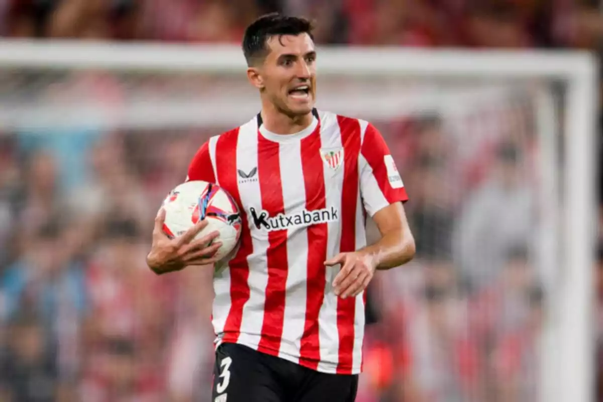 Un jugador de fútbol con uniforme rojo y blanco sostiene un balón en el campo.