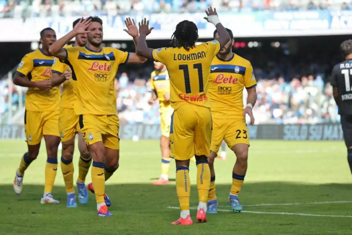 Jugadores de fútbol celebran un gol en el campo, vistiendo uniformes amarillos con detalles azules, mientras el público observa desde las gradas.