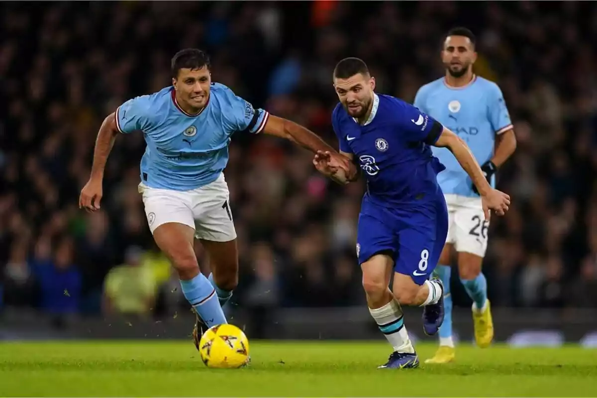 Dos jugadores de fútbol compiten por el balón en un partido, uno con uniforme celeste y el otro con uniforme azul, mientras un tercer jugador observa desde el fondo.