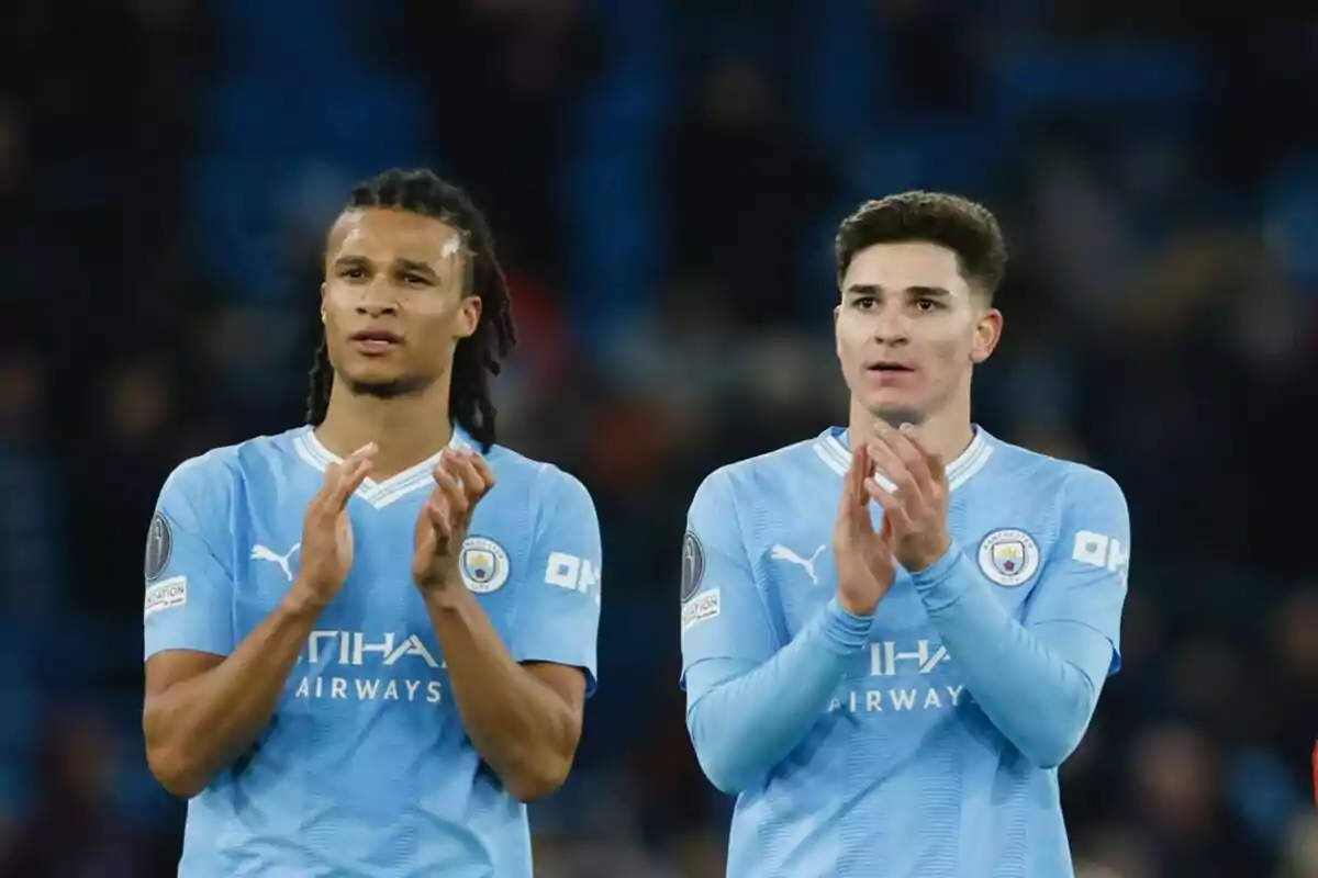 Dos jugadores de fútbol con uniforme azul claro aplauden en el campo.