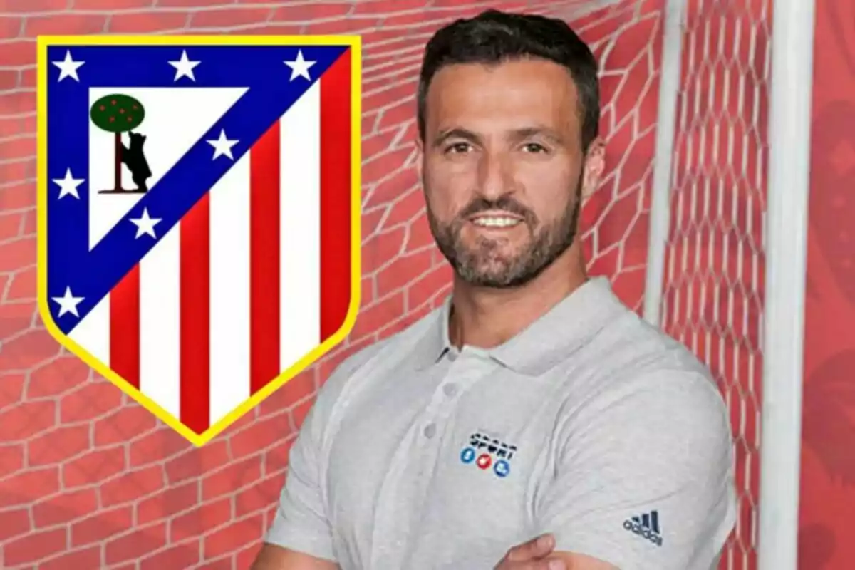 A man with a beard and a gray t-jersey poses in front of an Atlético de Madrid crest against a soccer net background.