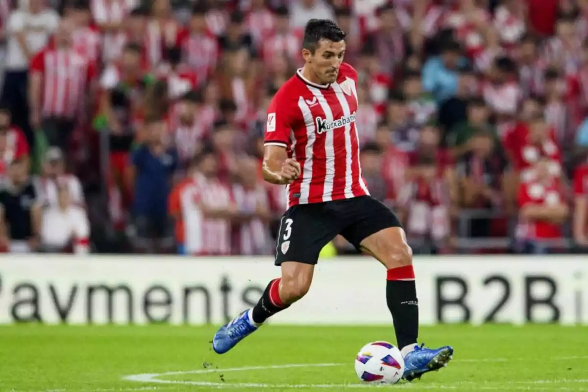 Un jugador de fútbol con uniforme rojo y blanco controla el balón en un estadio lleno de espectadores.