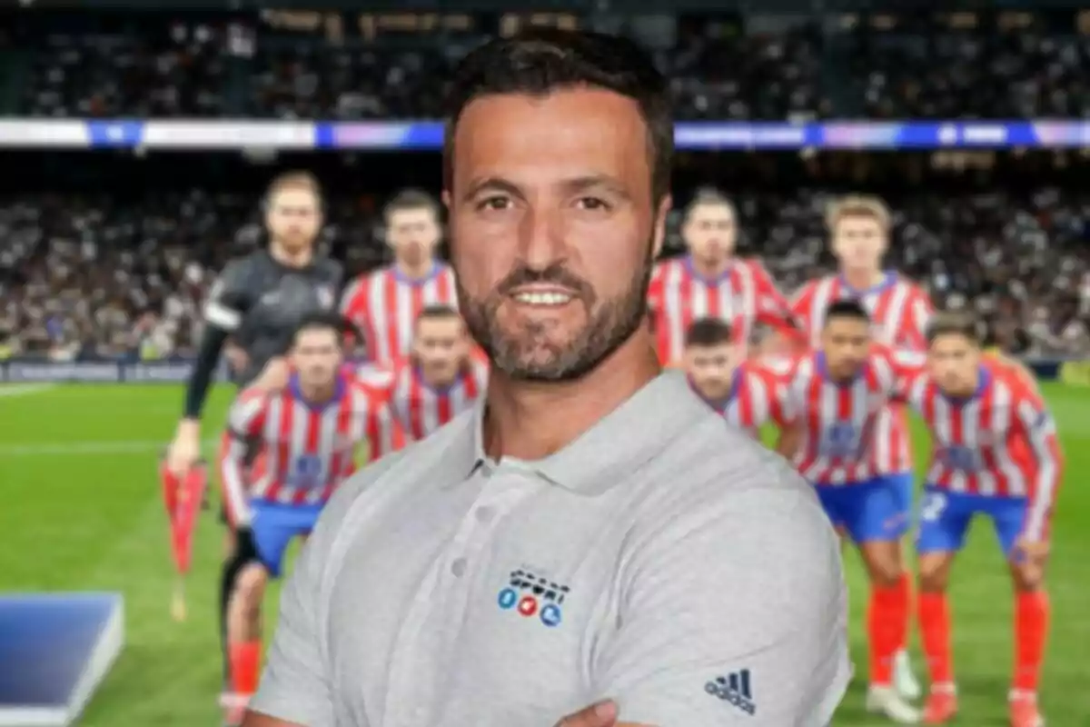 A man with a beard and a gray t-jersey is standing in front of a soccer team with red and white striped uniforms in a stadium full of spectators.