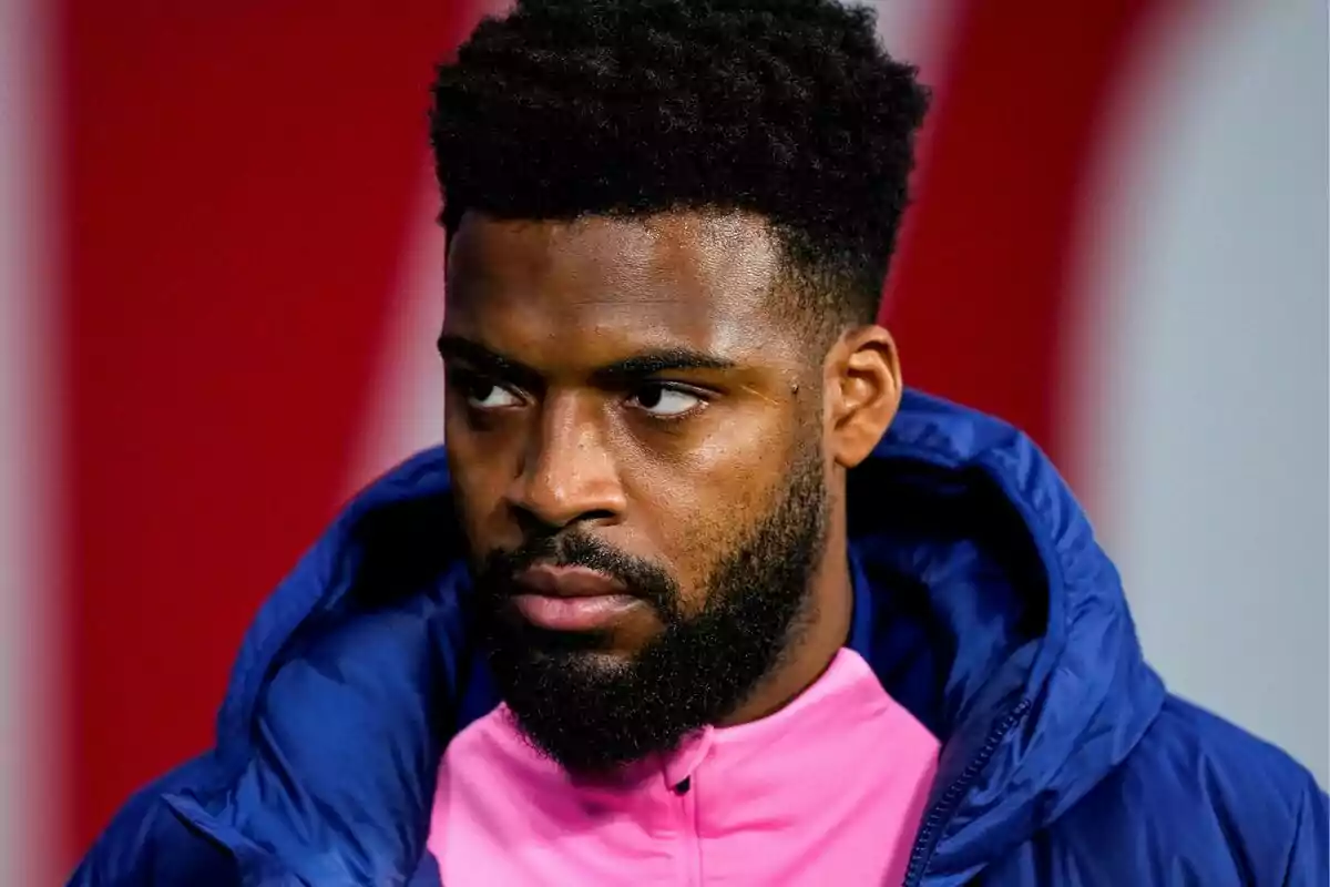 A man with a beard and curly hair is wearing a blue jacket and a pink t-jersey while looking to the side.