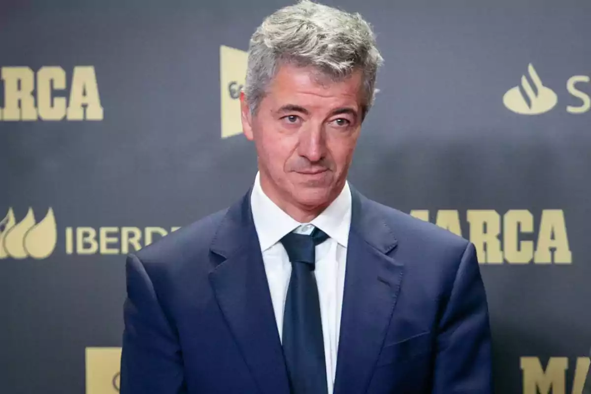 Hombre de cabello canoso con traje y corbata en un evento con fondo de logotipos de marcas.