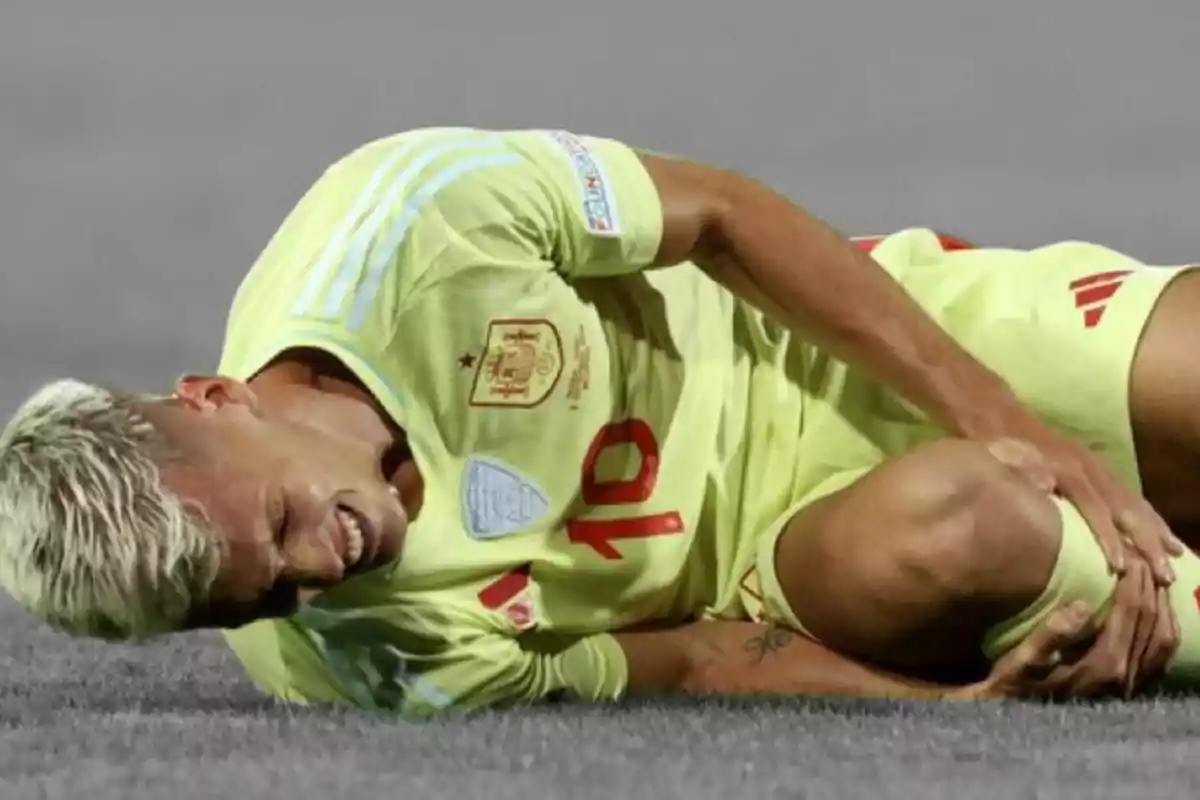 Jugador de fútbol con uniforme amarillo tendido en el suelo y sujetándose la pierna con expresión de dolor.