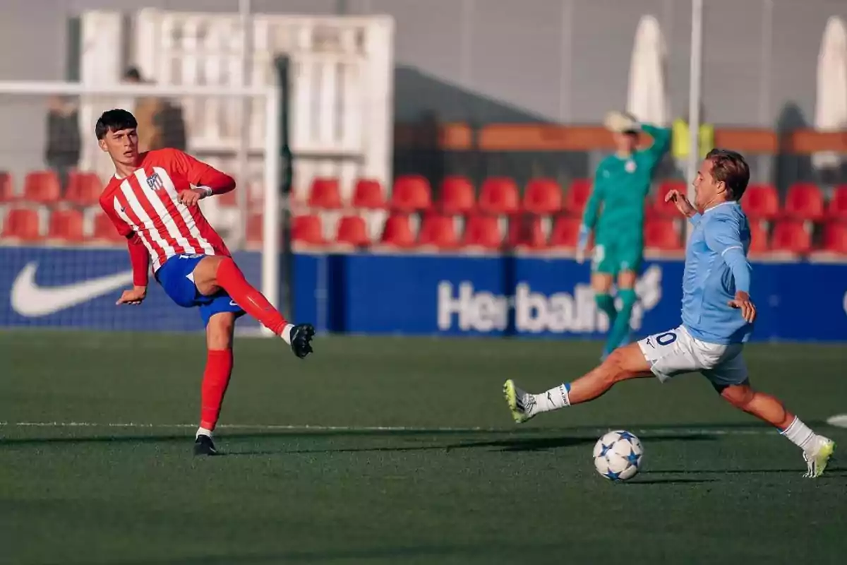 Dos jugadores de fútbol compiten por el balón en un campo, uno con uniforme rojo y blanco y el otro con uniforme azul claro.