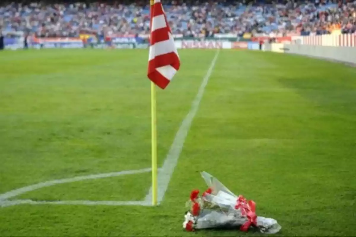 Un ramo de flores colocado en la esquina de un campo de fútbol junto a una bandera de esquina roja y blanca, con una multitud de espectadores en las gradas al fondo.