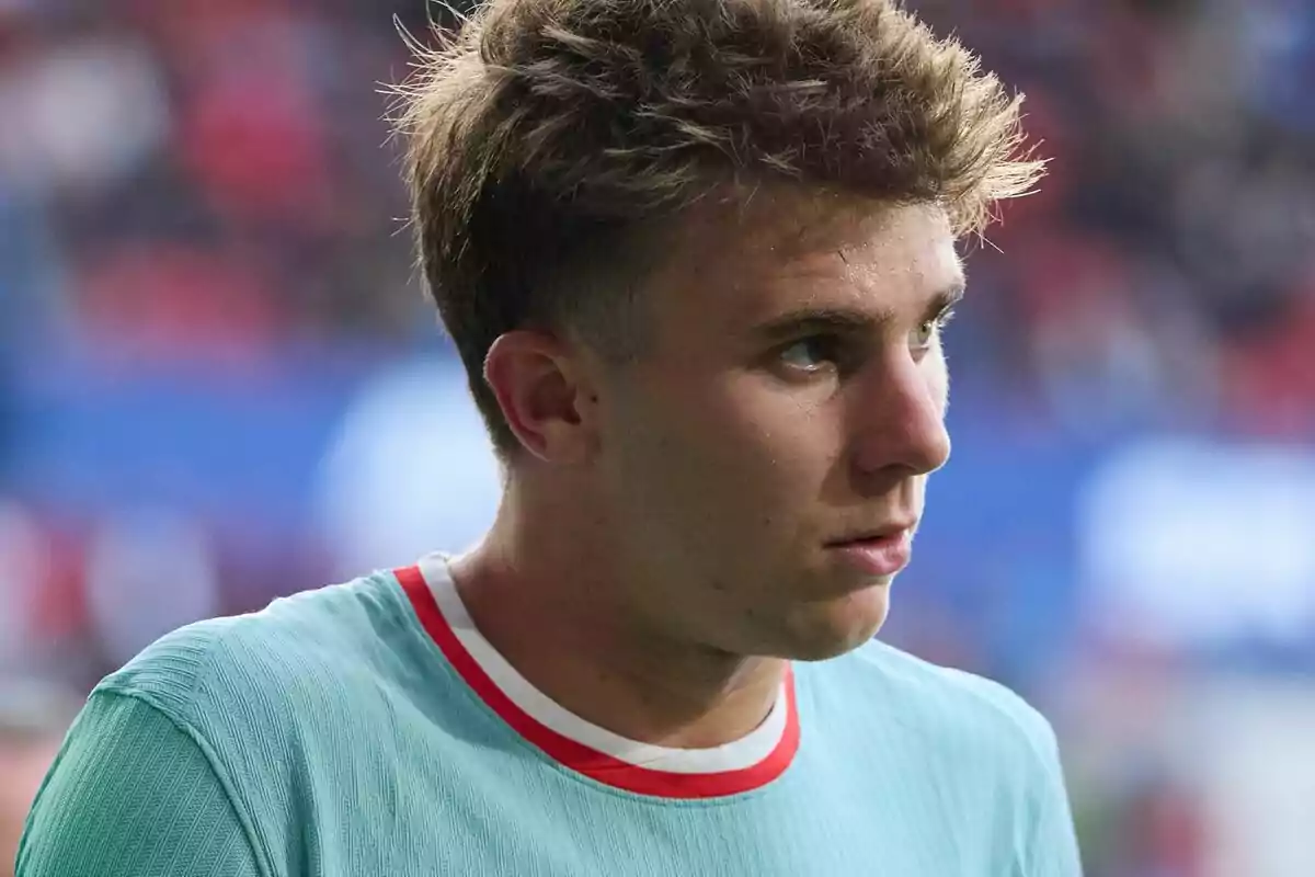 Un joven con camiseta deportiva de color verde claro y detalles rojos mira hacia un lado en un entorno de estadio.