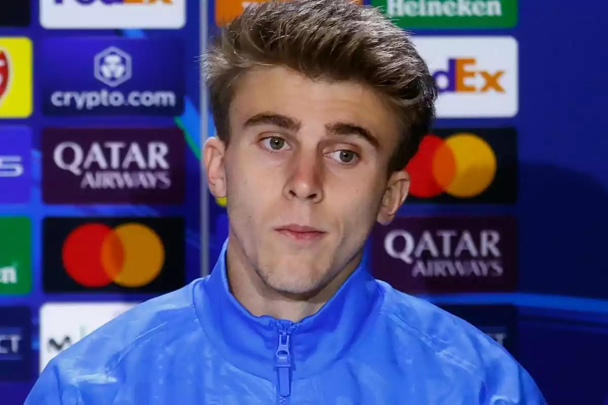 Un joven con chaqueta azul en una conferencia de prensa con logotipos de patrocinadores de fondo.