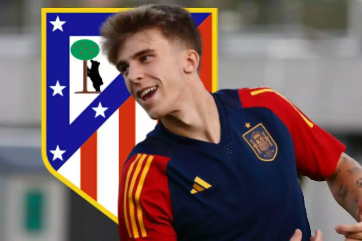 Un jugador de fútbol con la camiseta de la selección española sonríe con el escudo del Atlético de Madrid de fondo.