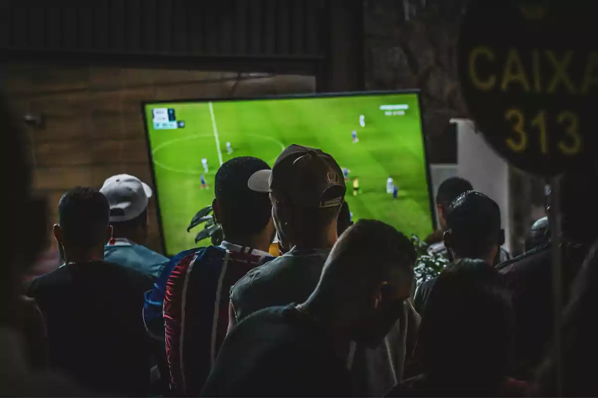 Un grupo de personas observa un partido de fútbol en una pantalla grande dentro de un lugar cerrado