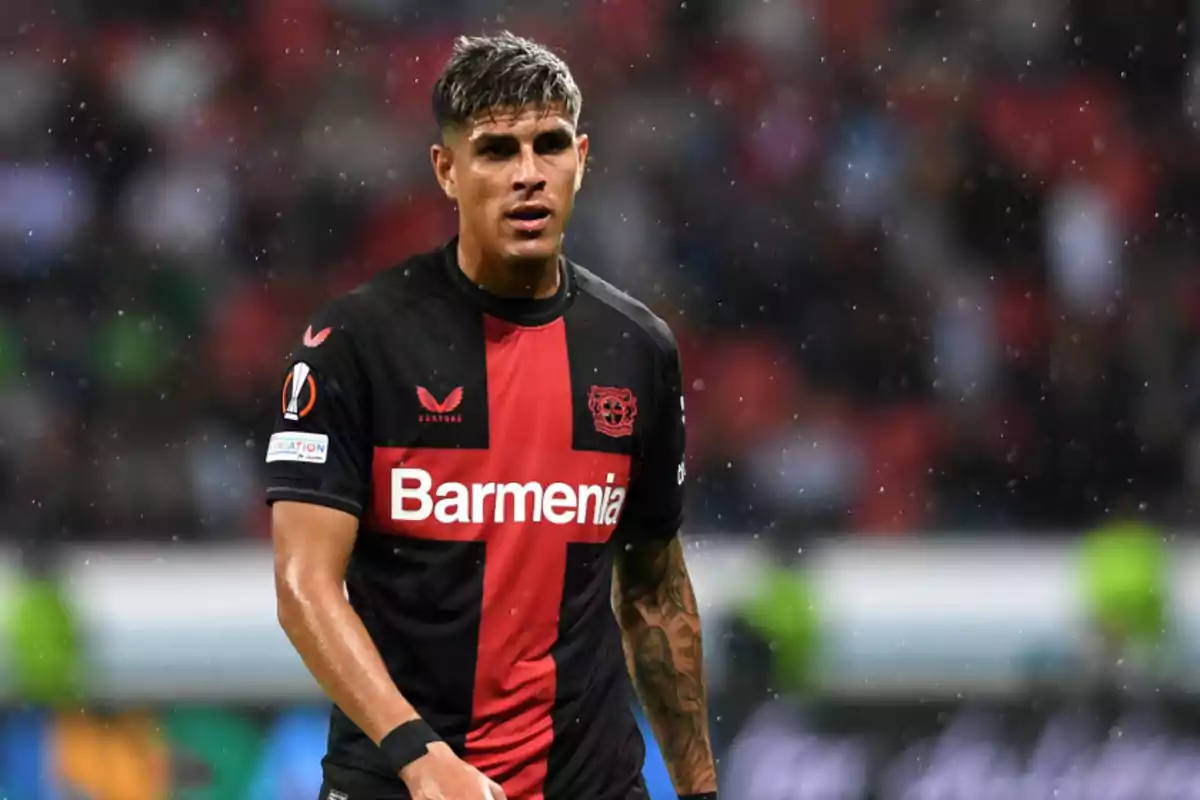 Jugador de fútbol con camiseta negra y roja del Bayer Leverkusen en el campo bajo la lluvia.