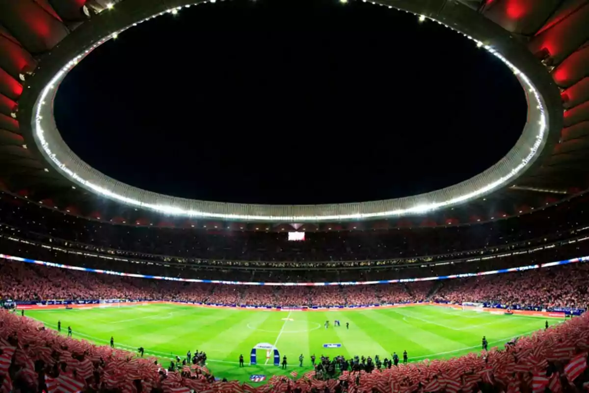 Estadio de fútbol iluminado durante un partido nocturno con aficionados en las gradas.