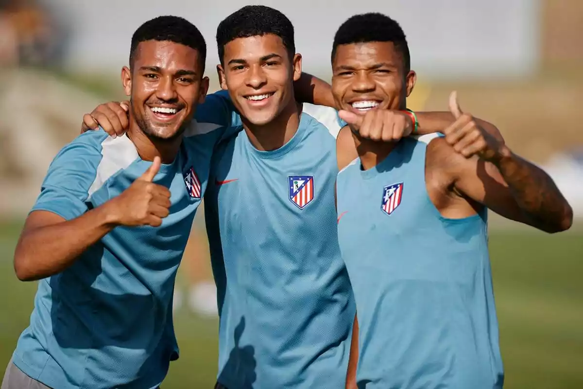 Tres jugadores de fútbol sonríen y levantan el pulgar mientras posan para una foto, todos llevan camisetas de entrenamiento del Atlético de Madrid.