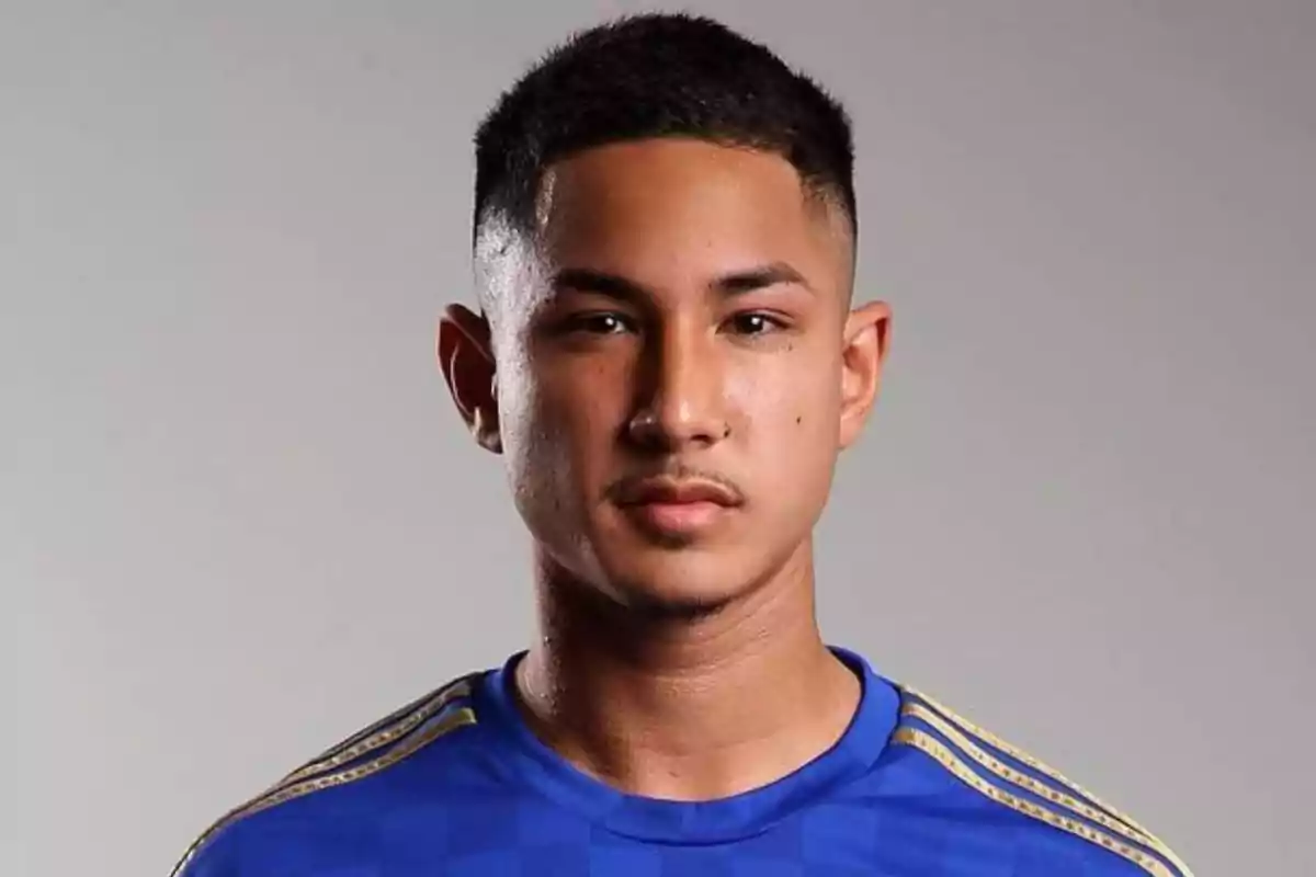 Un hombre joven con cabello corto y oscuro, vistiendo una camiseta azul con detalles dorados, posando frente a un fondo gris.