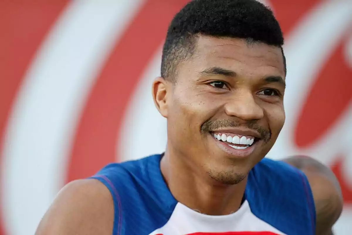 Un hombre sonriente con una camiseta deportiva azul y blanca, con un fondo desenfocado de color rojo y blanco.