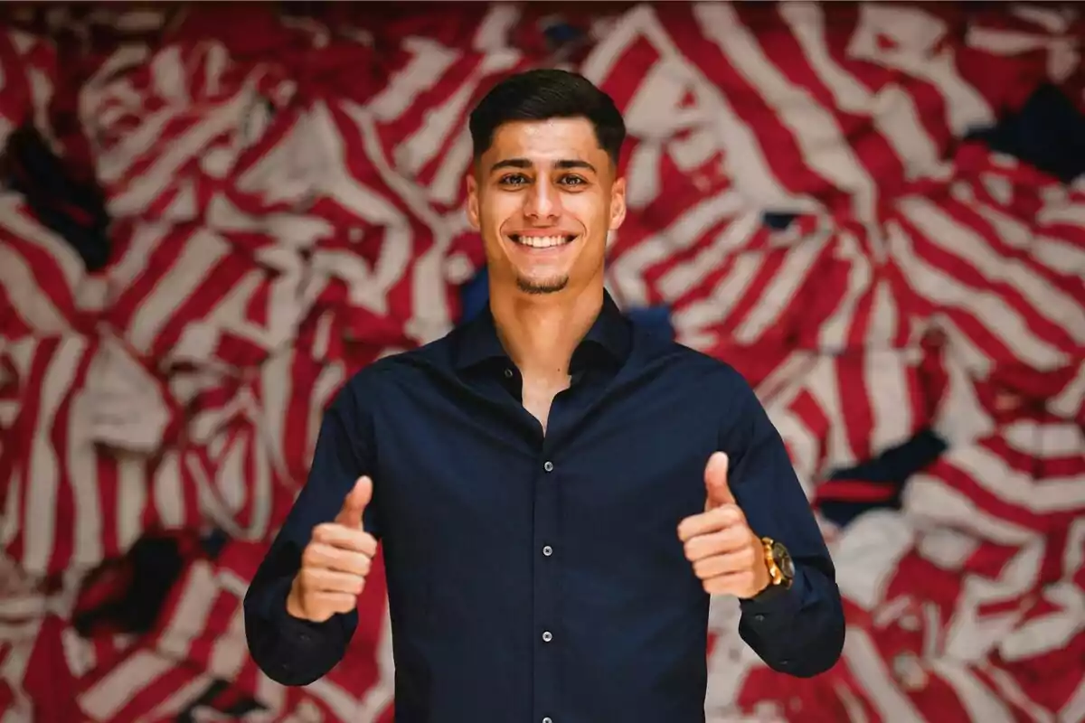 Un hombre joven con camisa oscura sonríe y muestra ambos pulgares hacia arriba, con un fondo de camisetas a rayas rojas y blancas.