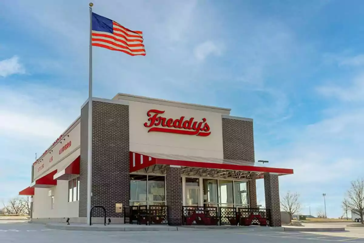 Restaurante Freddy's con fachada de ladrillo y toldos rojos, bandera de Estados Unidos ondeando y cielo despejado Restaurante Freddy's con fachada de ladrillo y toldos rojos, bandera de Estados Unidos ondeando y cielo despejado