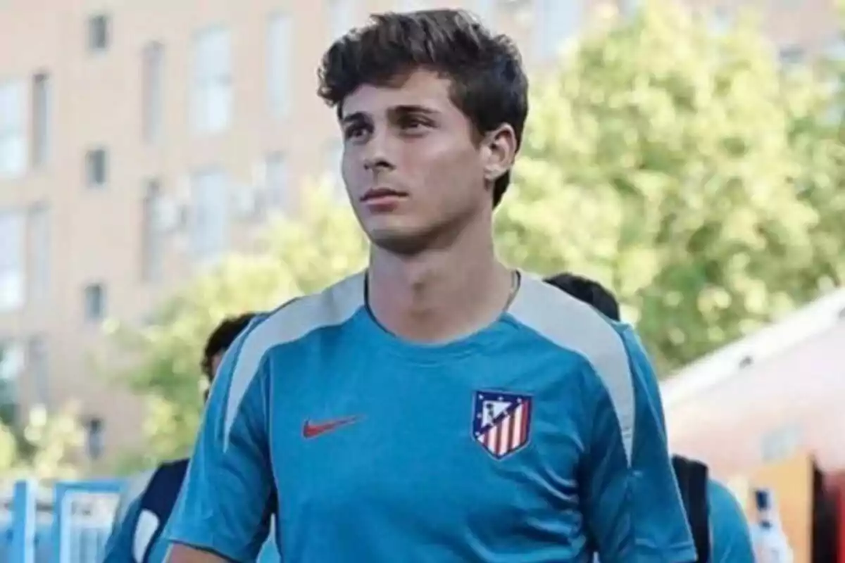 Un joven con una camiseta azul del Atlético de Madrid está de pie al aire libre con un edificio y árboles de fondo.