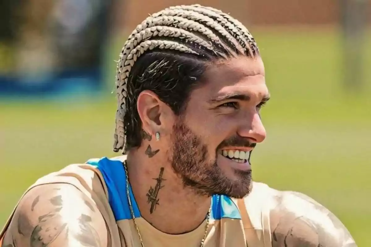 Un hombre con trenzas rubias y tatuajes en el cuello sonríe mientras está al aire libre.