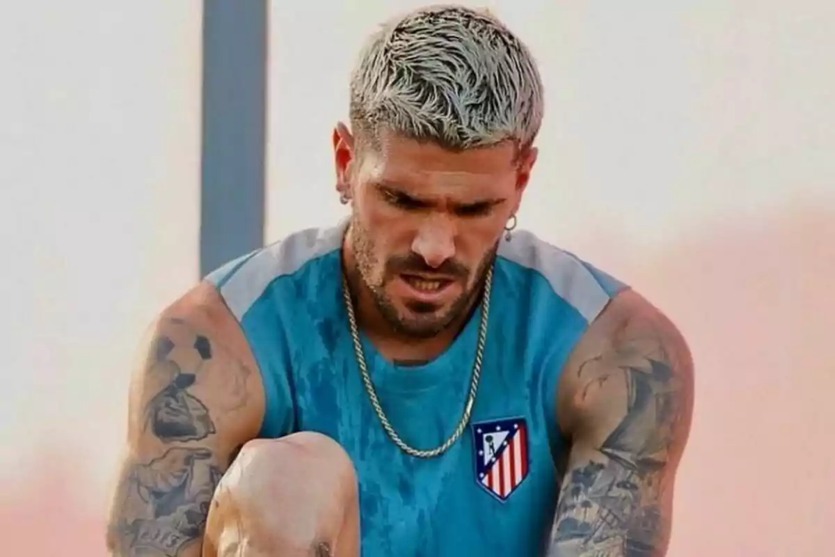 Un hombre con cabello rubio platinado y tatuajes en los brazos, vistiendo una camiseta sin mangas azul con el escudo del Atlético de Madrid, parece estar concentrado o cansado.