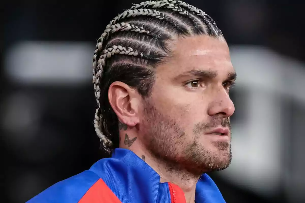 Un hombre con trenzas y tatuajes en el cuello, vistiendo una chaqueta azul y roja.