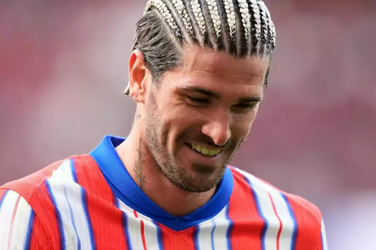 Hombre con trenzas y camiseta de rayas rojas y azules sonríe mientras mira hacia abajo.