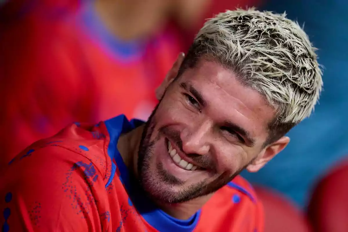 Un hombre con cabello rubio platinado y barba sonríe mientras viste una camiseta roja con detalles azules.