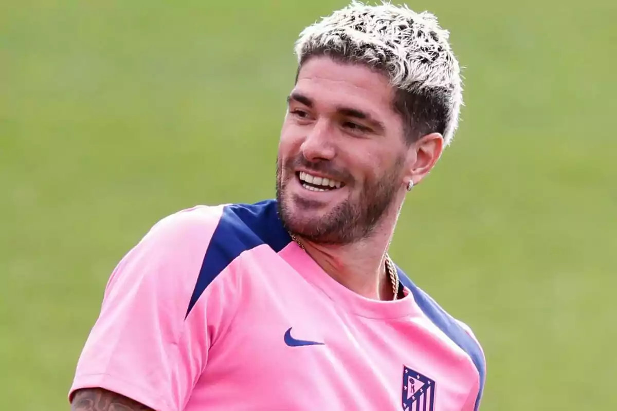 Un hombre con camiseta rosa y cabello teñido de rubio sonríe mientras mira hacia un lado.