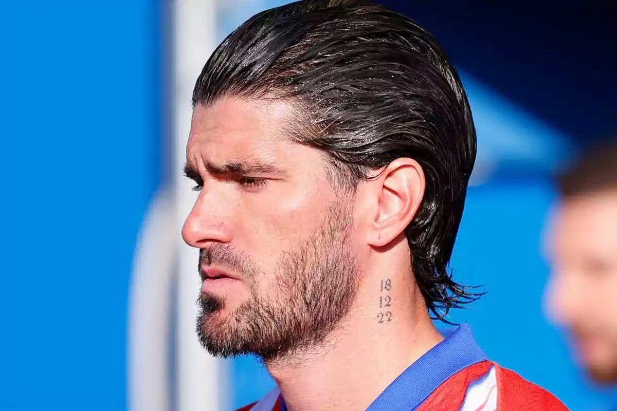 Man in profile with dark hair and beard, wearing a red and blue t-jersey and has a tattoo of numbers on his neck.