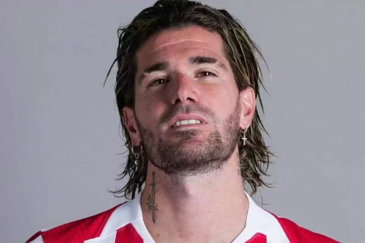 Hombre con cabello largo y barba usando una camiseta roja con blanco y aretes de cruz