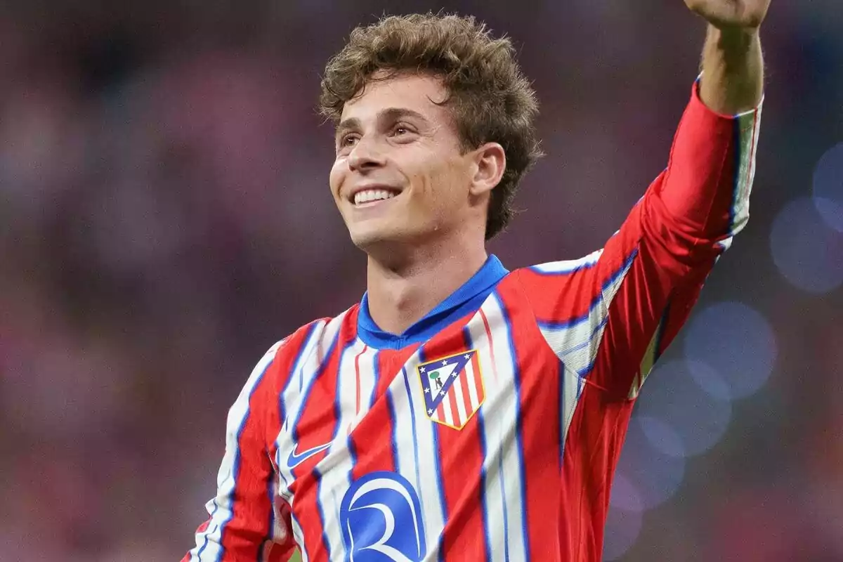 Jugador de fútbol con la camiseta del Atlético de Madrid sonriendo y levantando la mano.