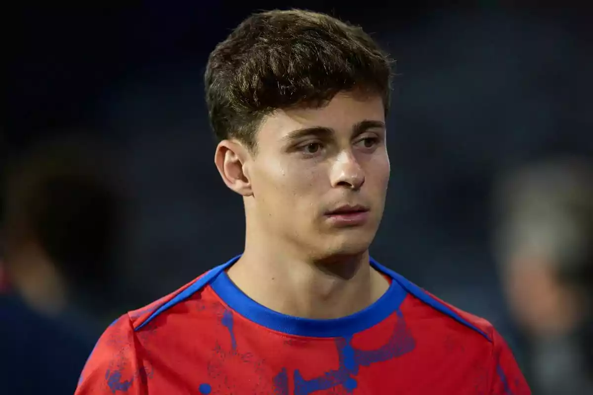 Un joven con camiseta roja y azul mira pensativo hacia un lado en un entorno oscuro.