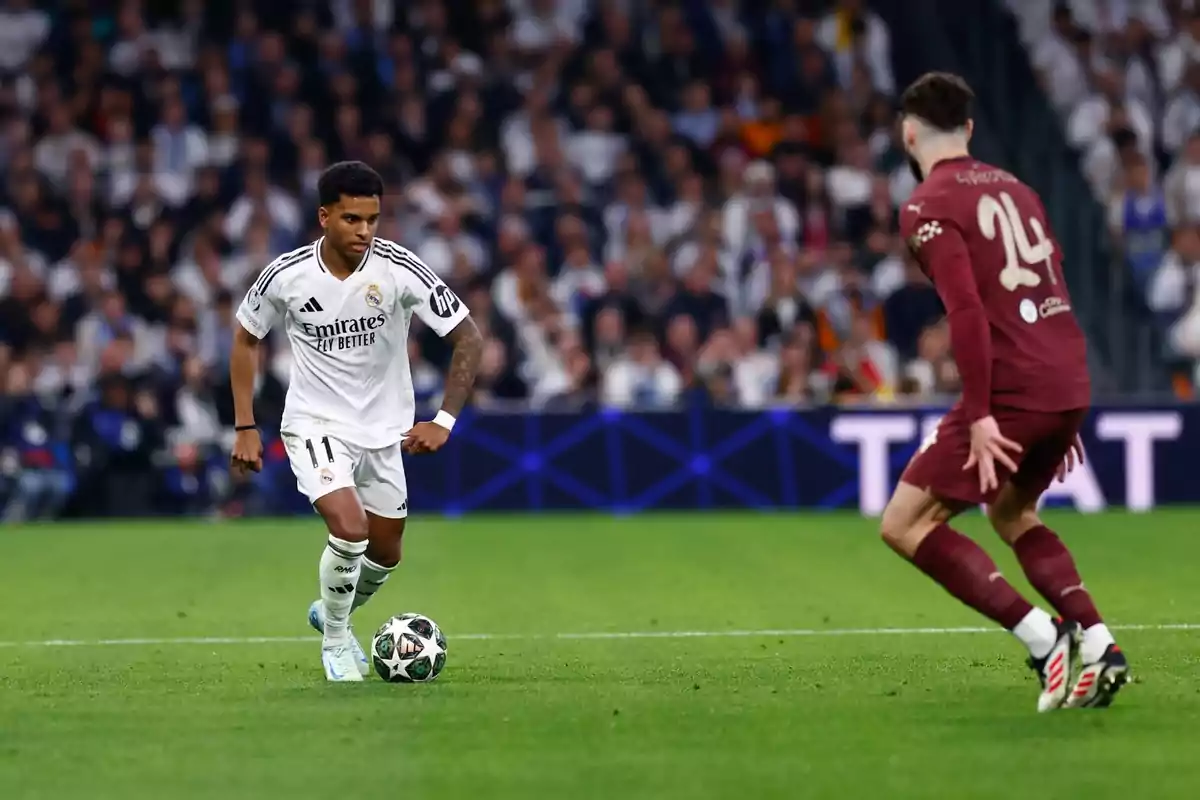 Un jugador de fútbol con uniforme blanco controla el balón mientras un oponente con uniforme marrón se acerca en un estadio lleno de espectadores.