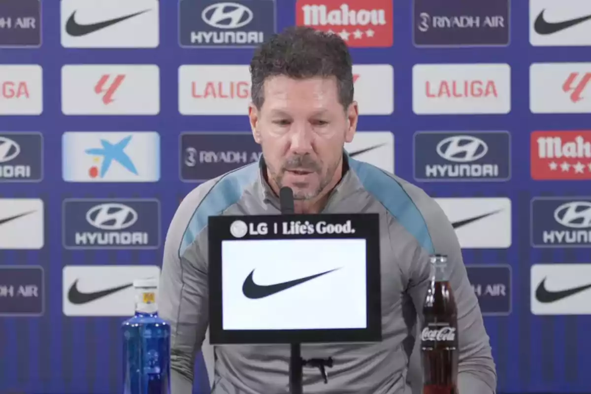 Un hombre en una conferencia de prensa con logotipos de patrocinadores en el fondo y botellas de agua y refresco en la mesa.