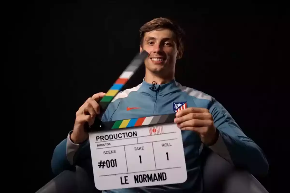 Un hombre sonriente con una chaqueta deportiva azul y el logo de un equipo de fútbol sostiene una claqueta de producción cinematográfica.