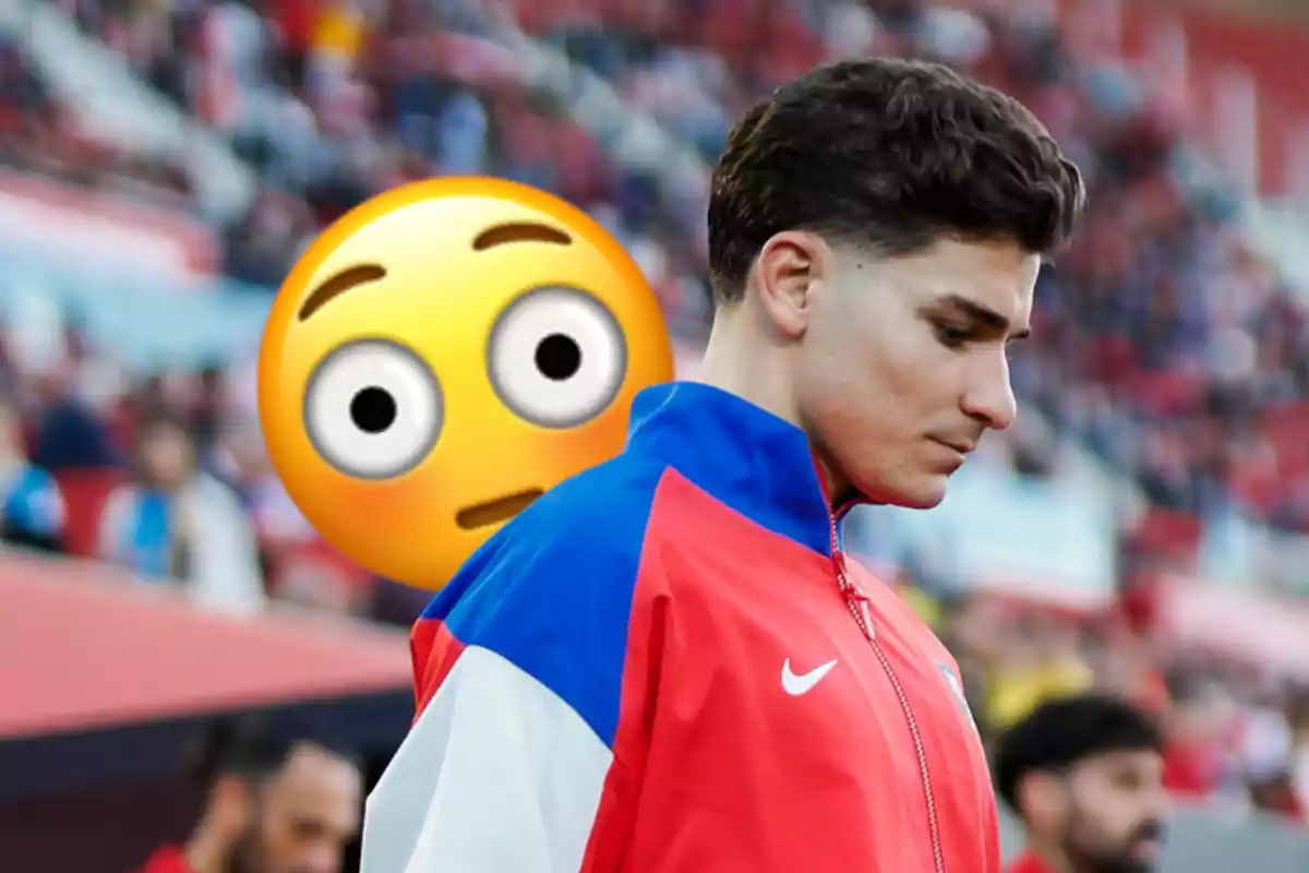 Un hombre con chaqueta deportiva roja y azul está de perfil en un estadio, con un emoji sorprendido detrás de él.