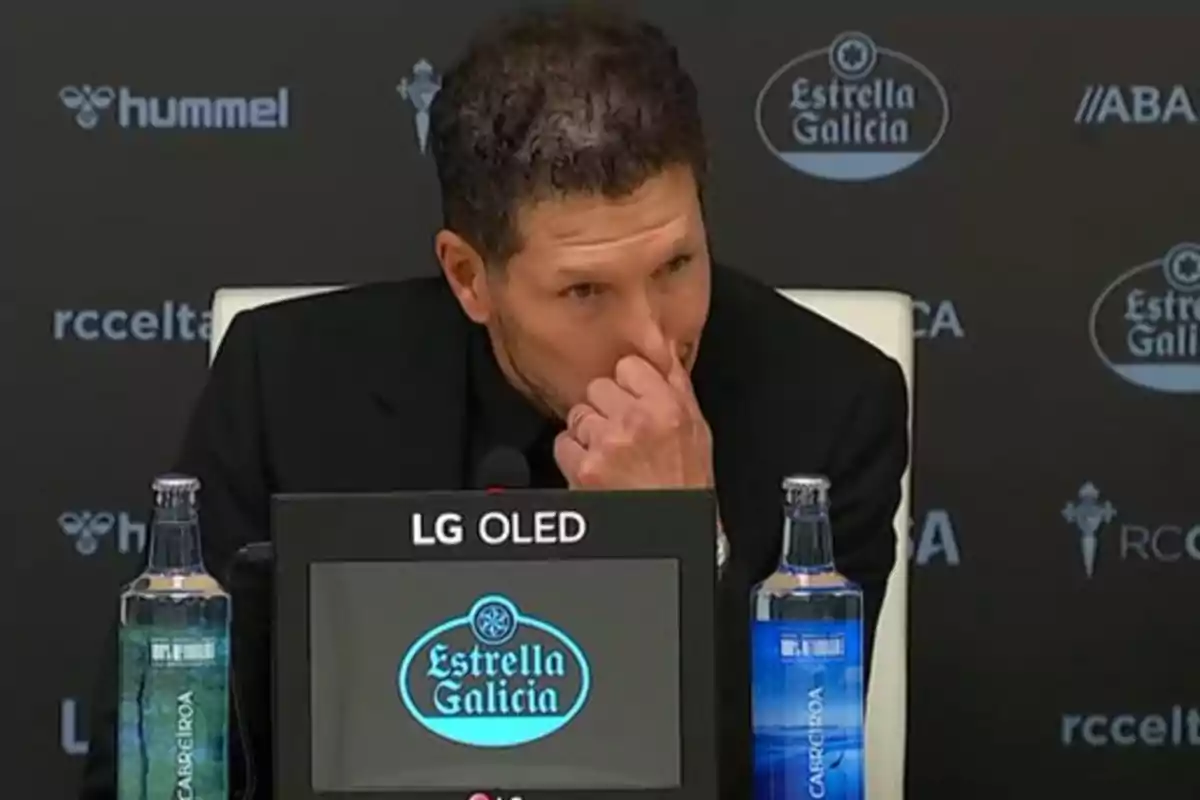 Un hombre en una conferencia de prensa con una pantalla que muestra el logo de Estrella Galicia y botellas de agua a los lados.