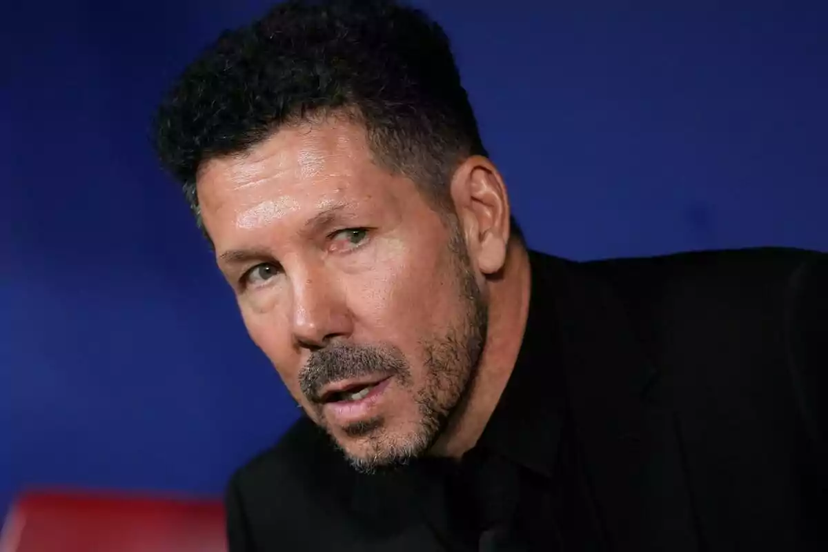 Hombre con barba y cabello corto, vestido de negro, en un fondo azul.