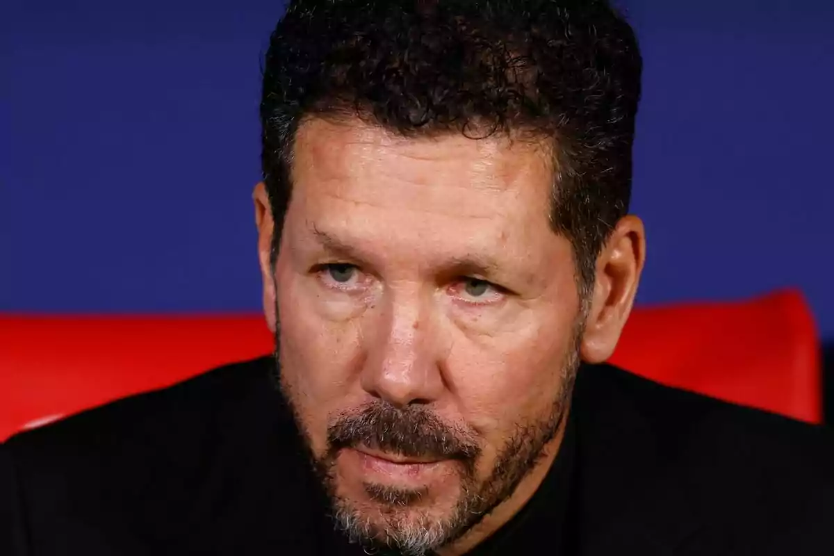 Un hombre con barba y cabello rizado mira hacia adelante con expresión seria.