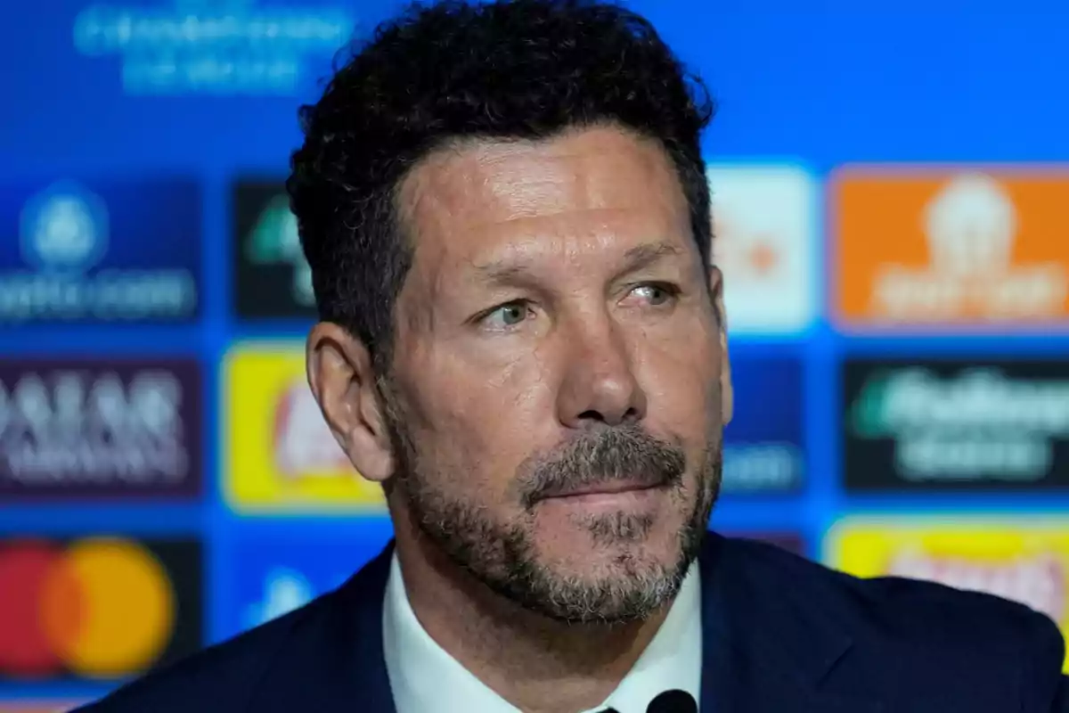 Un hombre con barba y cabello oscuro en una conferencia de prensa con un fondo azul y logotipos.