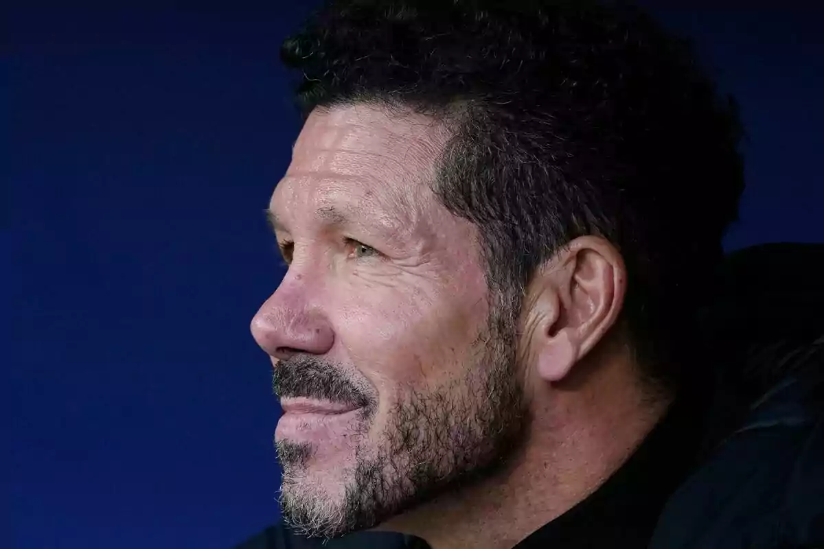 A man in profile with a beard and dark hair, looking forward with a thoughtful expression, against a blue background.