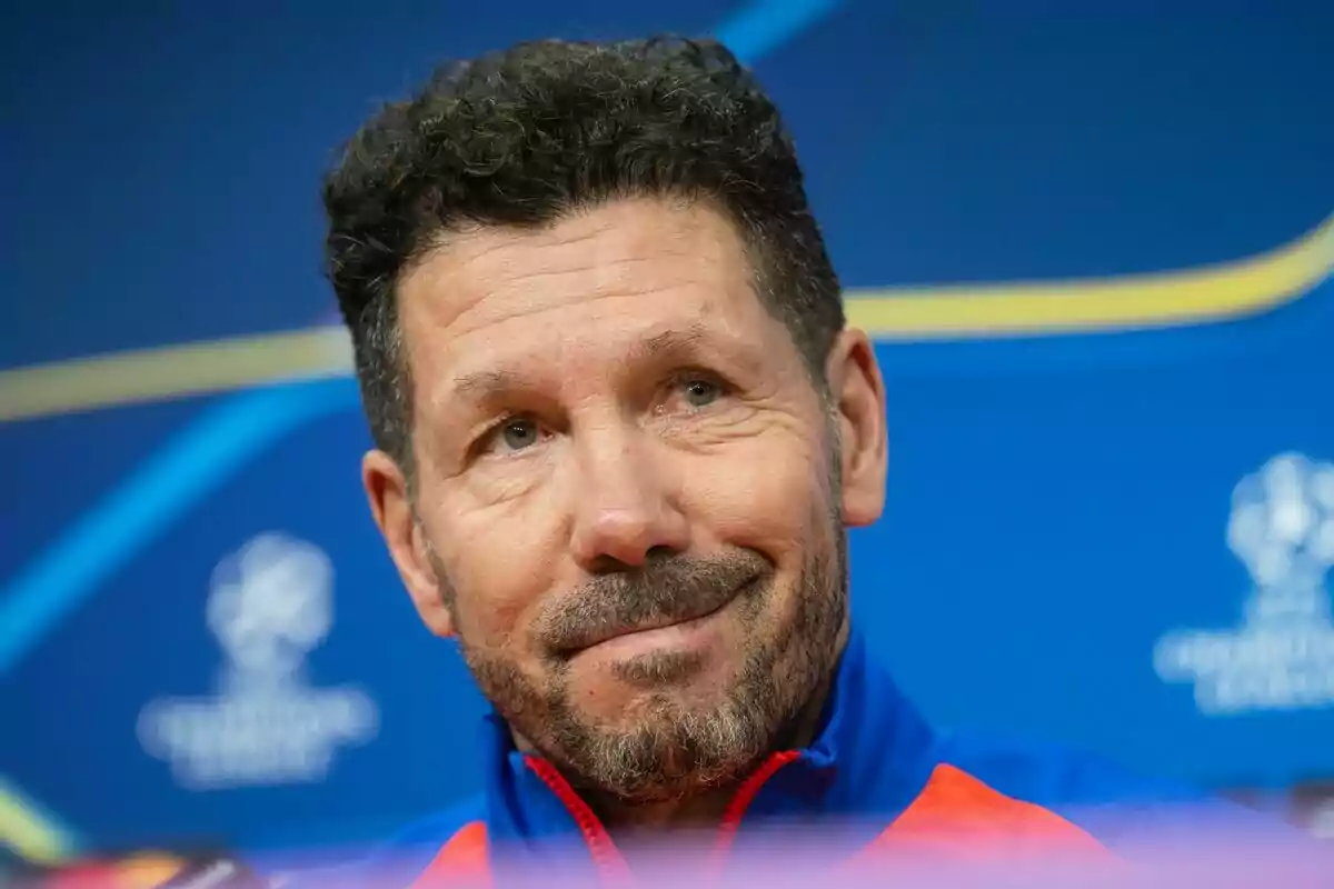 Un hombre con barba y cabello rizado sonríe ligeramente mientras está en una conferencia de prensa con un fondo azul.