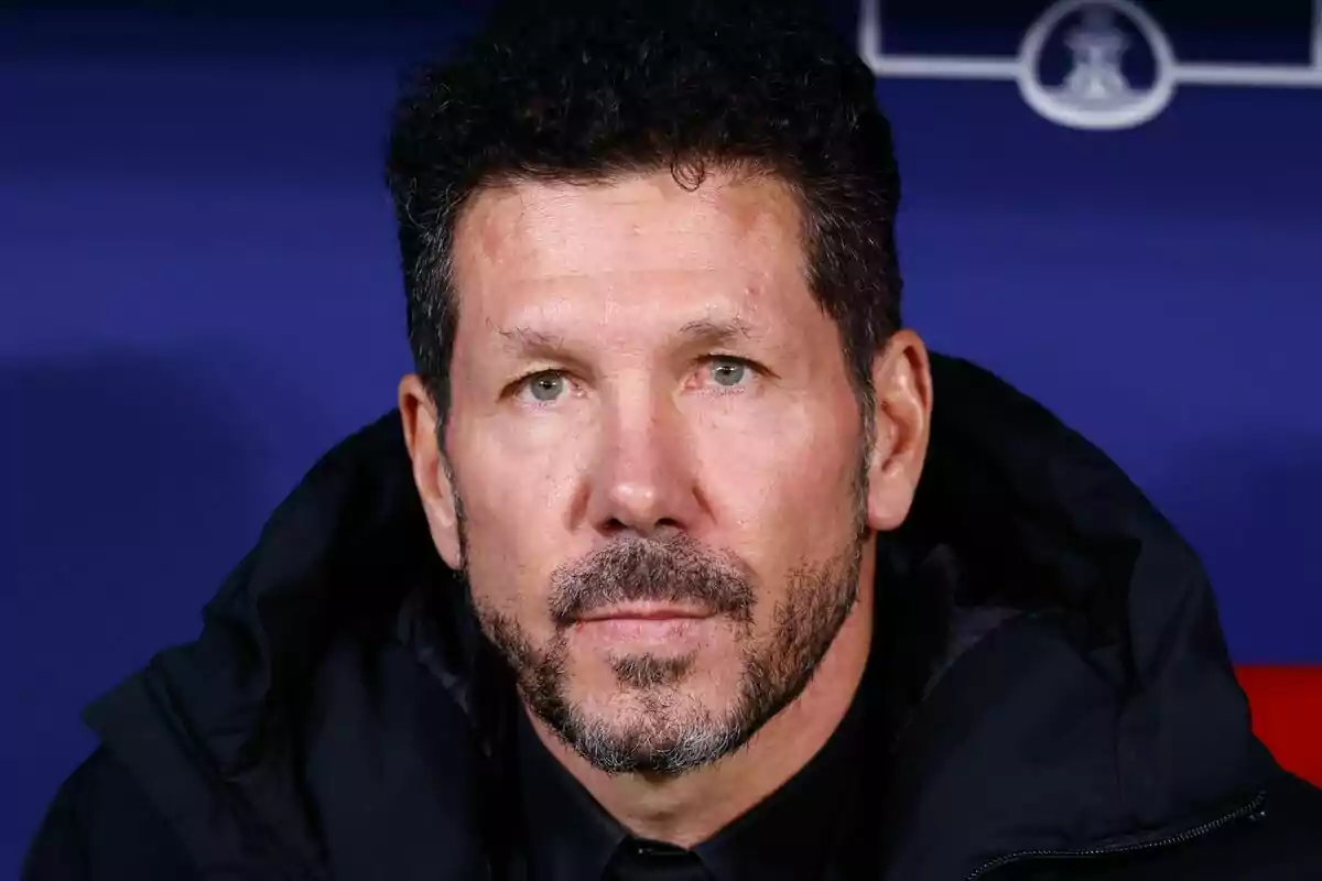 Un hombre con barba y cabello oscuro mira hacia adelante con expresión seria, vistiendo una chaqueta negra, con un fondo azul oscuro.