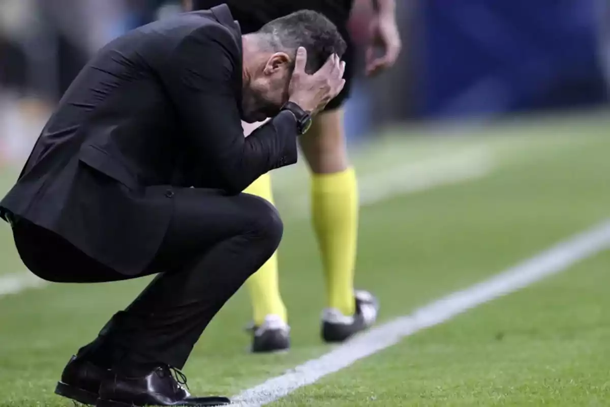 Un hombre en traje negro se agacha en el campo de fútbol con las manos en la cabeza mostrando frustración mientras un árbitro está de pie cerca.