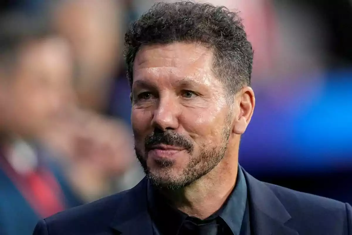 Hombre con barba y cabello rizado, vestido con traje oscuro, sonriendo.