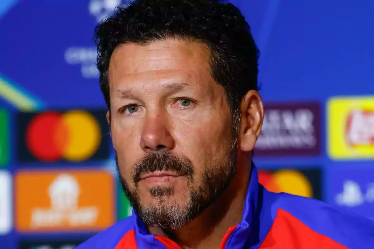 A man with a beard and dark hair at a press conference with a background of colorful logos.